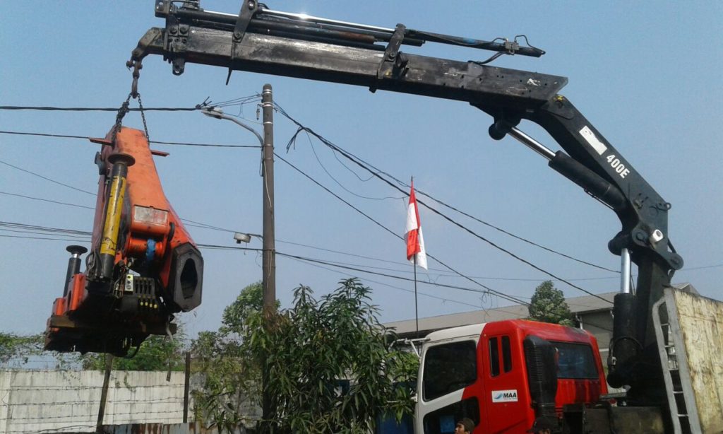 Truck crane MAA mengangkat unit alat berat berwarna oranye dengan sling di area kerja.