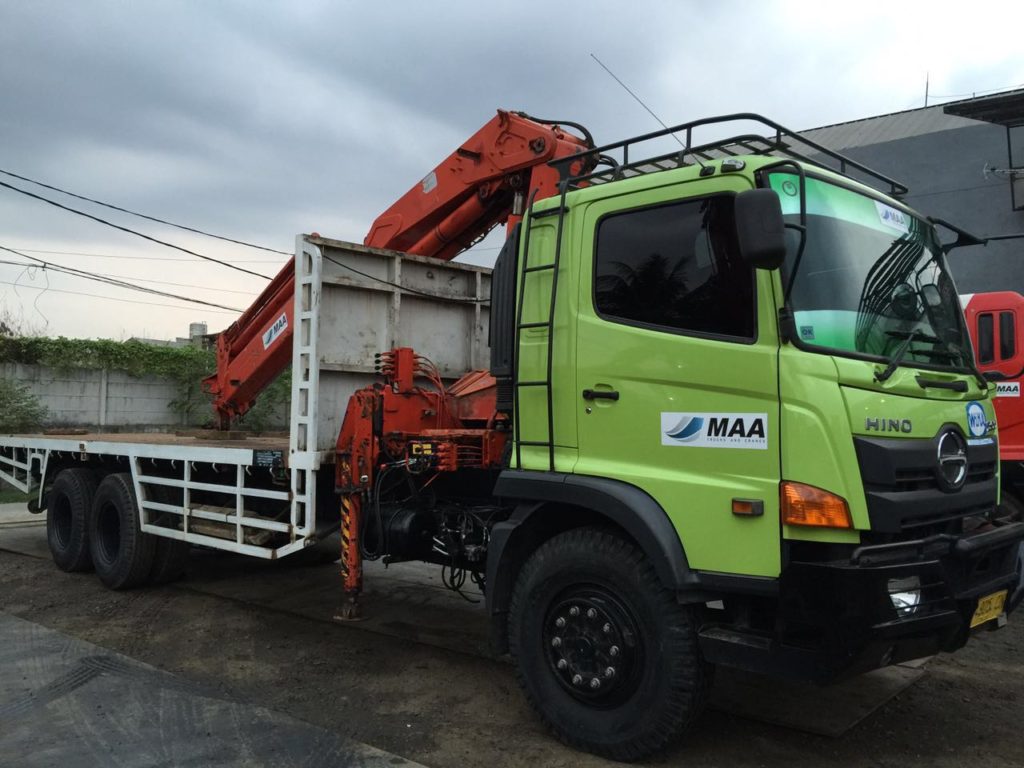 Truck crane Hino berlivery MAA dengan boom terlipat di atas bak muatan, siap dikirim ke lokasi proyek.