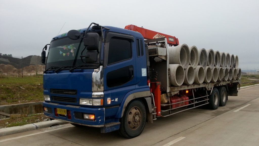 Truck crane biru mengangkut gorong-gorong beton di jalan menuju lokasi proyek.