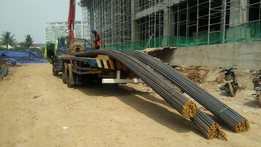 Pekerja menurunkan ikatan besi beton dari truck crane di area konstruksi bangunan bertingkat.