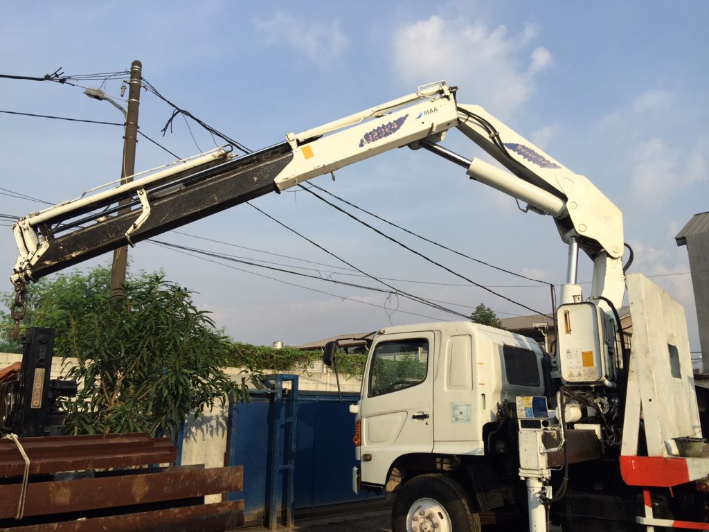 Mobile crane knuckle boom putih pada posisi terbuka di atas truck, di samping tumpukan material baja.